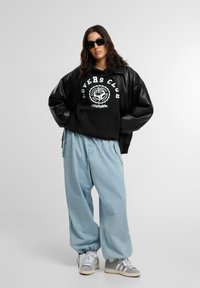 Juodas džemperis su „LOVERS CLUB“ grafika, derinamas su šviesiai mėlynomis plačiomis kelnėmis. Juodas odinis bomber tipo švarkas, pilki sportbačiai ir saulės akiniai.