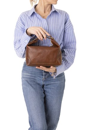 Donna con camicia a righe blu e jeans che tiene una borsa in pelle marrone con tracolla corta, che guarda di lato.