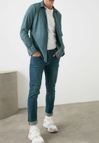 Camisa de denim azul claro sobre una camiseta blanca, combinada con jeans azul oscuro y zapatillas blancas. La camisa tiene un corte holgado y detalles de botones.