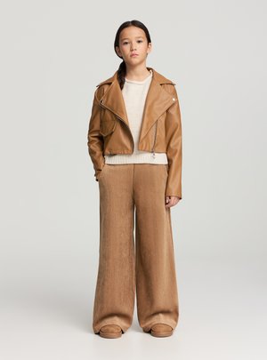 Giacca corta in pelle marrone, maglione beige, pantaloni marroni a gamba larga e calzature beige. Texture liscia e design aderente con dettagli a cerniera.