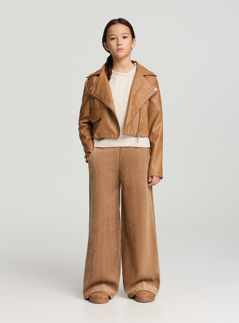 Giacca corta in pelle marrone, maglione beige, pantaloni marroni a gamba larga e calzature beige. Texture liscia e design aderente con dettagli a cerniera.