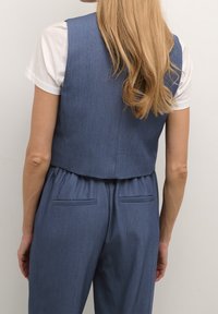 Niebieska dżinsowa kamizelka o krótkim kroju, z gładką fakturą i dwoma tylnymi kieszeniami, noszona na białej koszulce.