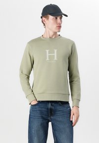 Sudadera de color verde oliva claro con cuello redondo, que presenta un logo blanco en forma de "H" y el texto "HACKETT". Combinada con jeans azules, con las manos en los bolsillos.