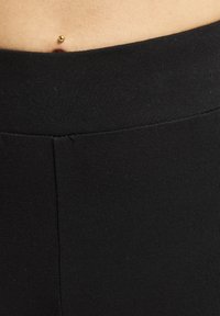Sort strækbar talje i leggings, der har en glat strikket struktur, og en lille synlig navlepiercing over taljen.