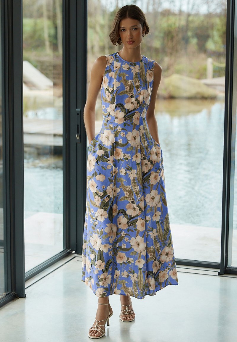 Femme debout à l'intérieur près de grandes fenêtres, portant une robe longue sans manches bleue à motifs floraux et des talons beiges à brides.