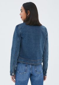 Veste en denim bleu foncé au design ajusté, à manches longues et avec des poignets à boutons. Présente un ourlet légèrement court et des détails de piqûre discrets.