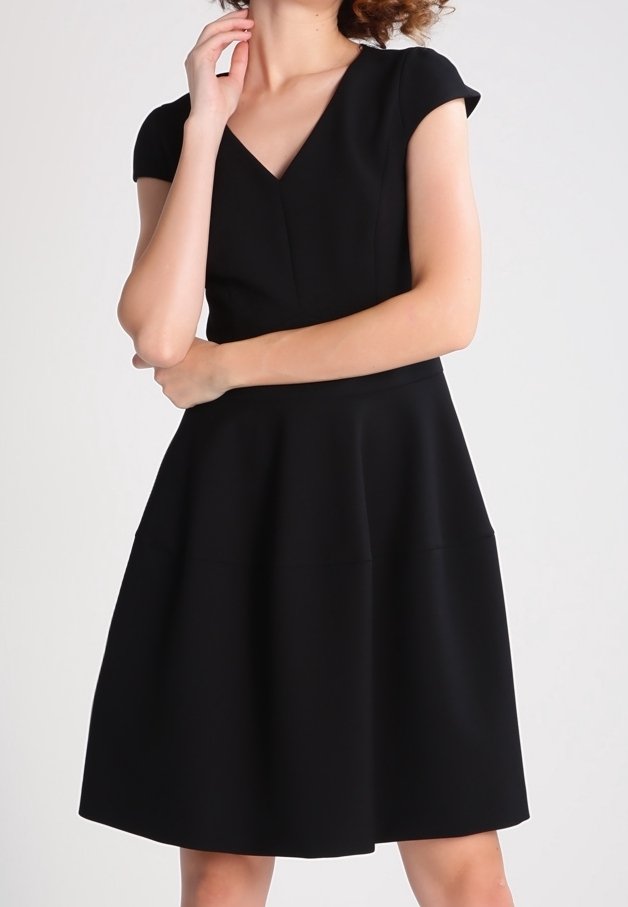 Femme portant une robe noire à manches courtes, col en V et jupe évasée, posant avec une main touchant son visage et l'autre bras croisé.