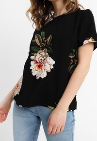 Black floral short-sleeve top featuring large white and beige flowers, green leaves, a loose fit, and a rounded neckline.