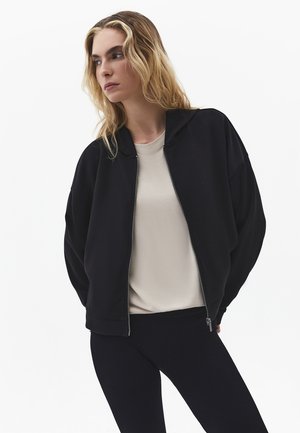 Vrouw met lang blond haar, gekleed in een zwarte jas met ritssluiting over een beige shirt en zwarte broek, kijkt opzij tegen een witte achtergrond.