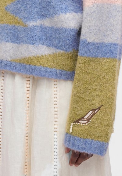 Manche de pull en maille douce rayée bleu, rose clair et vert, avec feuille marron brodée et détail perle sur une robe en dentelle blanche.