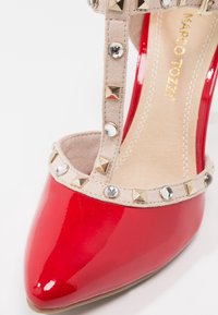 Escarpin en cuir verni rouge à talon haut avec un bout pointu. La sangle beigne est ornée de clous dorés et de strass. Texture lisse avec une finition brillante.