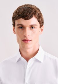 Wit overhemd met kraag en een gladde textuur, voorzien van een knoopsluiting aan de voorkant en een klassieke pasvorm, gedragen door een persoon met kort, golvend haar.