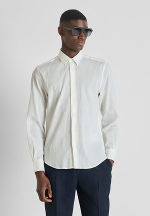 Uomo che indossa una camicia bianca a bottoni con trama, pantaloni scuri e grandi occhiali da sole neri, in posa davanti a uno sfondo grigio chiaro neutro.