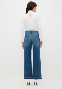 Biała bluzka z długim rękawem z wycięciem na plecach, w zestawie z niebieskimi jeansami o wysokim stanie i szerokich nogawkach, posiadającymi tylne kieszenie i postrzępiony mankiet.