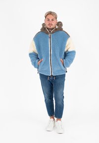 Fluffy blauwe en crèmekleurige jas met een capuchon, met een rits aan de voorkant en bruine accenten. Draagt lichtblauwe jeans en witte sneakers.