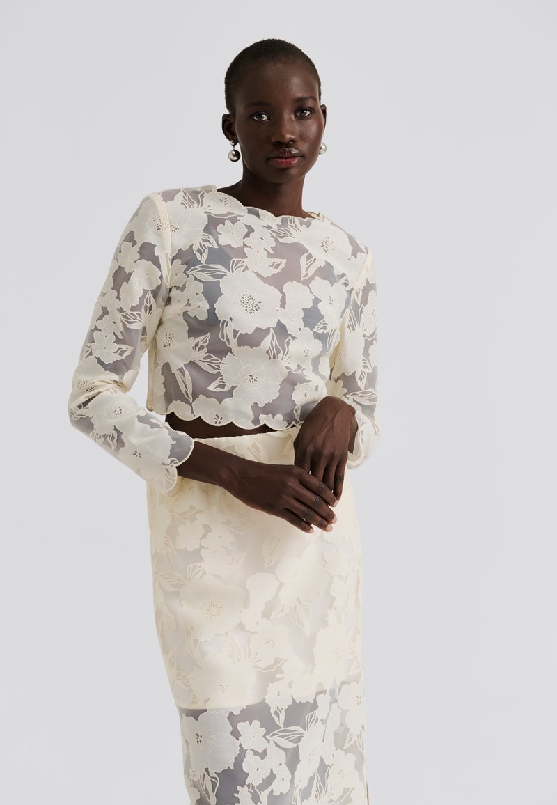 Csipkés kétdarabos öltözék, áttetsző virágmintás elefántcsontszínű felsővel, hosszú ujjal, és hozzá illő, testhezálló, ugyancsak elefántcsontszínű virágmintás szoknyával.