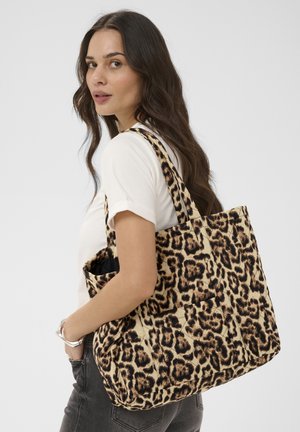 Frau mit langen dunklen Haaren, die ein weißes T-Shirt und Jeans trägt und eine große Tasche mit Leopardenmuster über der Schulter trägt.