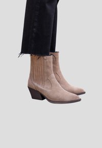 Bottines en daim beige avec bouts pointus et talons carrés, portées avec un jean noir court à ourlet effiloché, sur un fond neutre.
