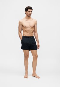 Homme torse nu aux cheveux courts et foncés, portant un short de bain noir, debout pieds nus sur un fond clair uni.