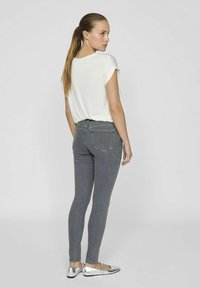 Jeans ajustados grises con detalles en los bolsillos, combinados con una blusa blanca de manga corta. La modelo lleva zapatos planos metálicos y está de espaldas a la cámara.