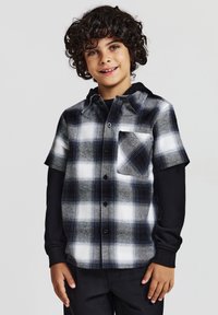 Camicia a maniche corte a quadri in bianco, nero e grigio. Presenta un colletto, una tasca sul petto ed è sovrapposta a una maglietta nera a maniche lunghe.