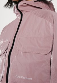 Light pink nylon jacket featuring a shiny finish, adjustable hood, black zipper, and front pocket. Brand name printed on the hem.