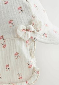 Baumwollmütze mit floralem Muster in sanftem Rosa auf weißem, strukturiertem Hintergrund. Mit Schleifenakzent und gerafftem Rand für zusätzlichen Charme.