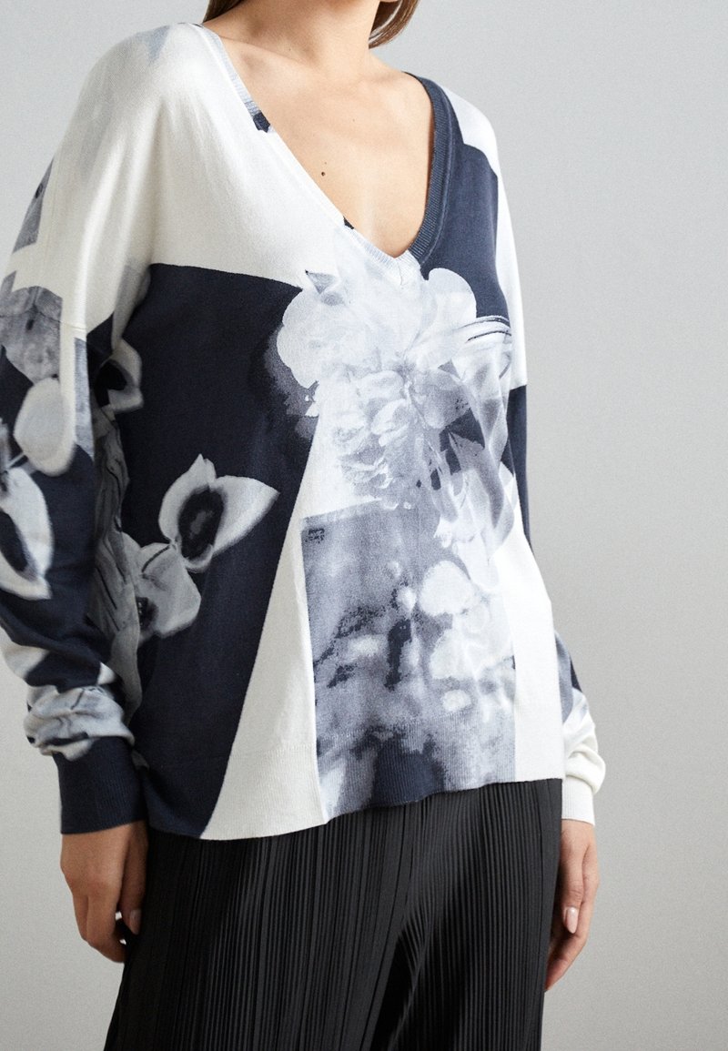 Pull à col en V blanc et marine, orné d'un design floral et d'une texture douce. Manches oversize et coupe décontractée. Associé à un pantalon plissé.