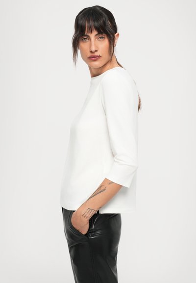 Woman with dark hair and bangs wearing a white long-sleeve top and black leather pants with one hand in pocket against a plain background.