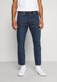 Tmavě modré džíny z denimu se rovnými nohavicemi, pěti kapsami a jemnými švy. Spárované s bílými teniskami a jednoduchým tričkem.