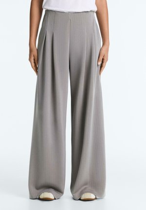 Kobieta nosząca jasno szare, szerokie spodnie w prążki z wysokim stanem i plisami, połączone z białymi płaskimi butami i włożonym białym topem.