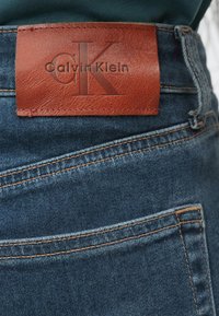 Tmavě modré džíny z denimu s hnědou koženou nášivkou na pasu s vyraženým nápisem "Calvin Klein" a logem "CK", zdůrazňující detaily stehu.