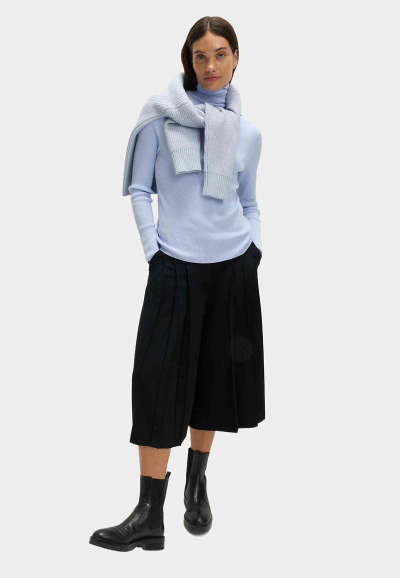 Frau trägt einen hellblauen Rollkragenpullover mit einem passenden Pullover über den Schultern, schwarze weit geschnittene 3/4-Hosen und schwarze Stiefeletten.