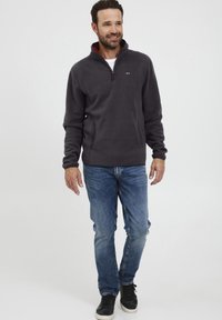 Pelēks vilnas džemperis ar augstu apkakli un sarkanu iekšpusi, ar sānu kabatām, valkāts ar zilām džinsām un melniem apaviem.