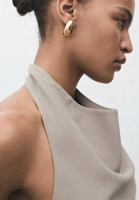 Haut à col halter beige en tissu lisse, associé à des boucles d'oreilles en or en forme d'ovale, mettant en avant un design minimaliste.
