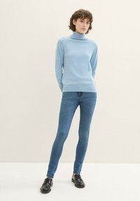 Suéter de cuello alto azul claro, textura acanalada, combinado con jeans ajustados de color azul y zapatos negros de cordones, de pie contra un fondo neutro.
