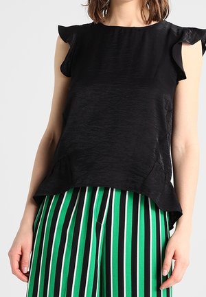 Kvinde iført en sort ærmeløs top med flæsekanter og bukser med lodrette grønne, hvide og sorte striber.