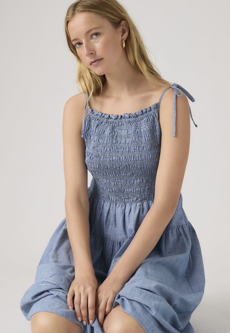 Jeune femme aux cheveux blonds portant une robe bleue smockée sans manches avec des bretelles à nouer, assise les mains reposant sur ses genoux.