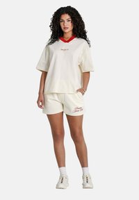 Completo in cotone color crema composto da una maglietta oversize a maniche corte con scollatura a V rossa, abbinato a pantaloni corti coordinati e sneakers bianche.
