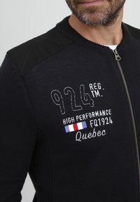 Jachetă tricotată neagră cu fermoar frontal, umeri texturați și broderie albă ce include numere și text, printre care "Quebec" și un design cu steag.