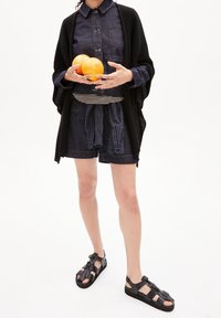 Jeansskjorta med knappar, marinblå shorts, svart stickad cardigan och svarta sandaler. Håller två apelsiner, neutral bakgrund.