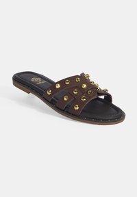Bruine sandaal met een platte zool, voorzien van drie brede banden versierd met gouden studs, een zwarte platte zool en een onderzijde van natuurlijk rubber met accentstiksels.