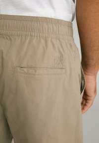Pantalons beiges avec une taille élastique, dotés d'une poche arrière et d'un détail de logo brodé. Texture lisse ; design ajusté.