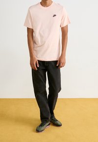 Jasnoróżowa bawełniana koszulka z czarnym logo Nike, zestawiona z czarnymi dżinsami i zielonymi sneakersami z purpurowymi akcentami, na żółtej podłodze.