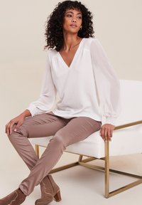 Weiße Bluse mit V-Ausschnitt und transparenten langen Ärmeln, kombiniert mit beigen, eng anliegenden Hosen und braunen Stiefeletten mit Blockabsatz.