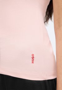 Világos rózsaszínű, testhez simuló póló puha anyagból, apró piros "HUGO" logóval az alsó oldalon. Sima textúra és ívelt szegély.