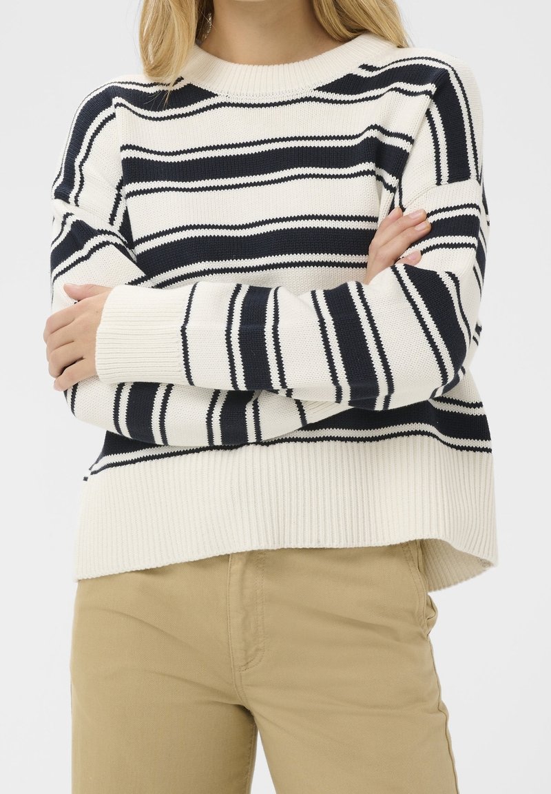 Personne portant un pull rayé noir et blanc avec les bras croisés et un pantalon beige, montrée du cou aux hanches sur un fond uni.