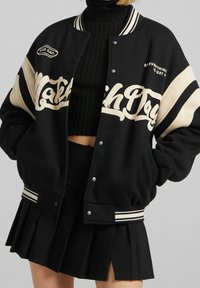Schwarze Collegejacke mit cremefarbenen Akzenten, Druckknöpfen und gestreiften Rippbündchen. Mit grafischem Text auf der Vorderseite und lockerer Passform.