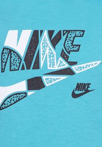 Tirkīza krāsas T-krekls ar izteiksmīgu, daudzkrāsainu Nike logotipu baltā, melnā un teksturētā dizainā, kā arī nelielu melnu svītru.