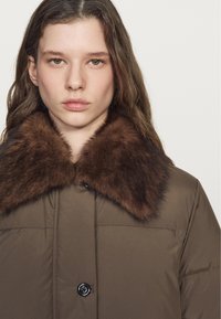 Giovane donna con capelli castani alle spalle che indossa un cappotto imbottito marrone con un grande colletto in pelliccia marrone scuro, sfondo neutro.